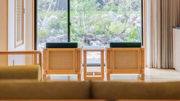 Una habitación con dos sillas de mimbre con cojines verdes, una pequeña mesa de madera y un ventanal corredero que da a un jardín con rocas y follaje.