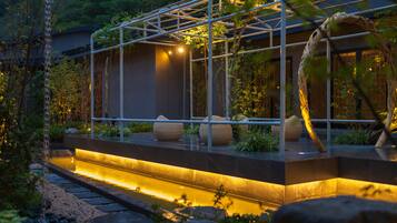 A modern outdoor seating area with a glass enclosure, a water feature, and surrounding greenery.
