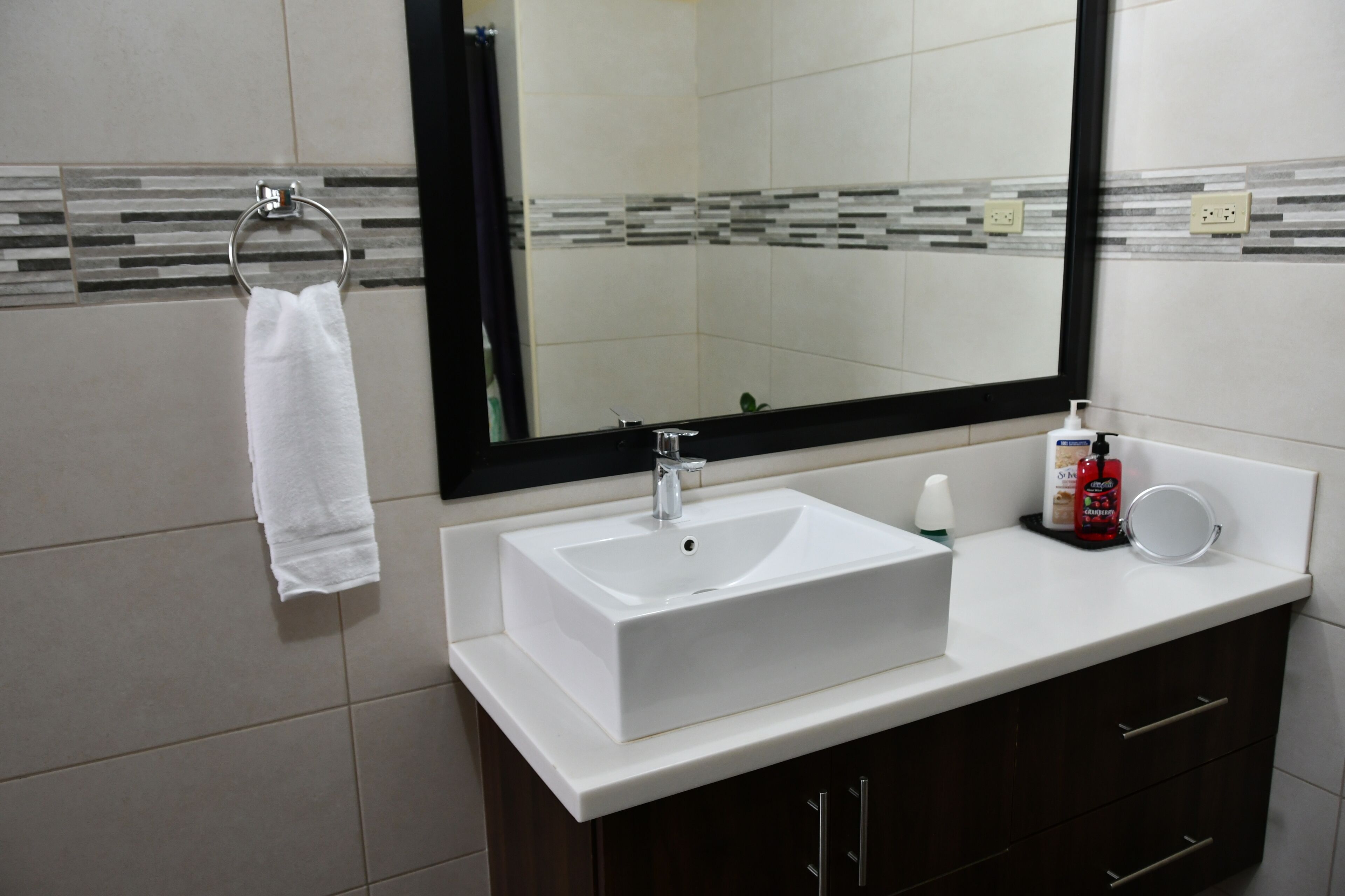 Un baño con un lavamanos blanco, un espejo, un aro para toallas y un dispensador de jabón.