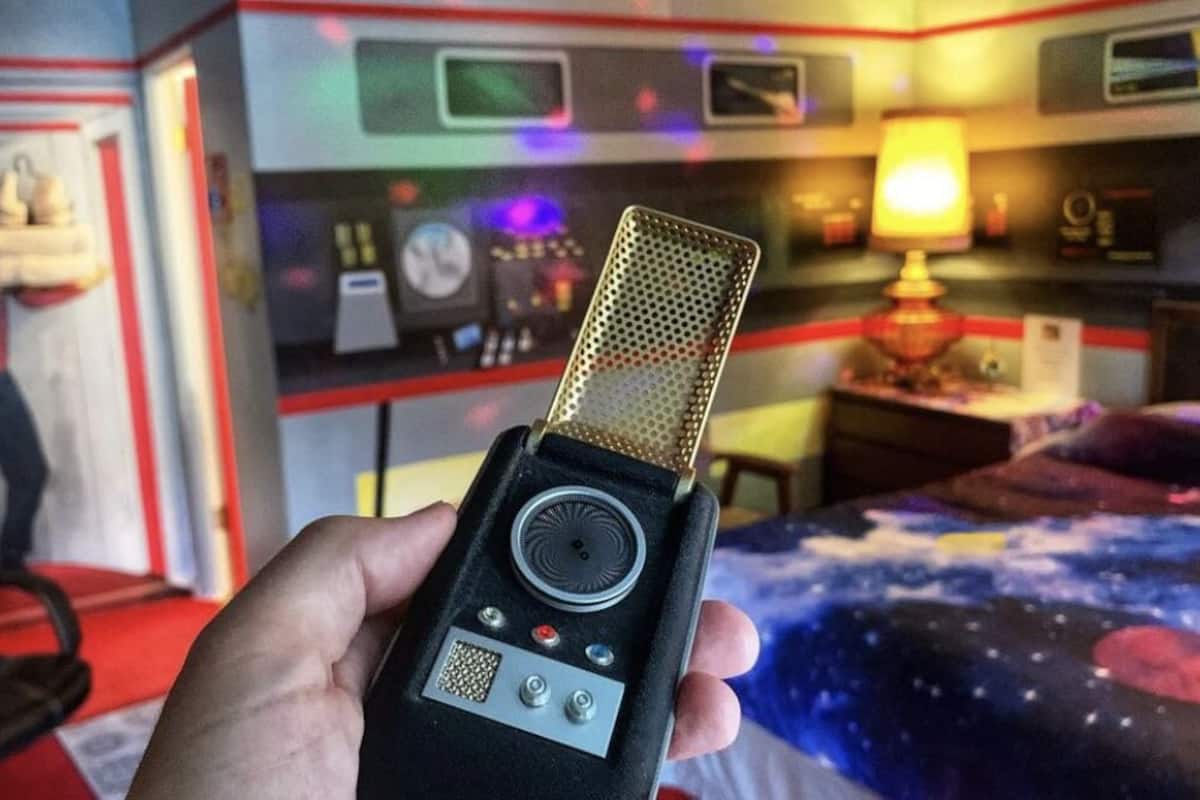 Eine Hand hält ein altes Mikrofon mit Bedienfeld, in einem Raum mit einem Bett, das mit einem Bettbezug im Galaxendesign ausgestattet ist, einem Schreibtisch mit Lampe und einer Wand mit einer Uhr und anderen Gegenständen.