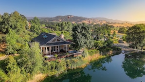 Una casa con patio que da a un lago tranquilo, rodeada de exuberante vegetación y colinas al fondo.
