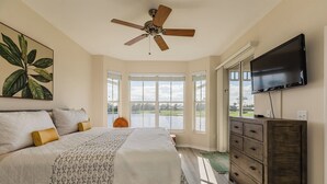 Une chambre à coucher avec un lit, un ventilateur de plafond, une télévision et de grandes fenêtres donnant sur une étendue d’eau.