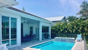Une piscine avec une terrasse carrelée, un patio couvert avec des chaises, et une vue sur une maison dotée de grandes fenêtres.