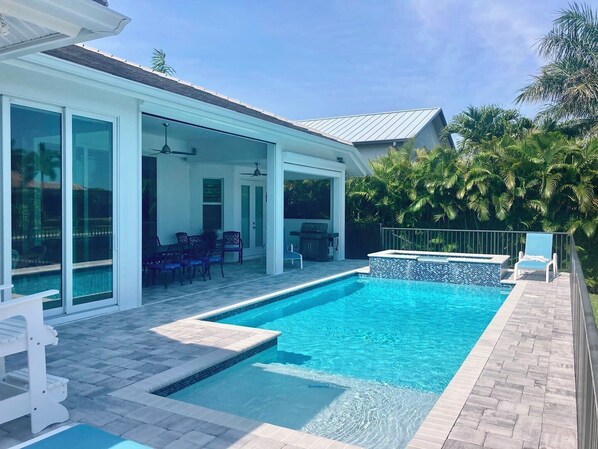 Une piscine avec une terrasse carrelée, un patio couvert avec des chaises, et une vue sur une maison dotée de grandes fenêtres.
