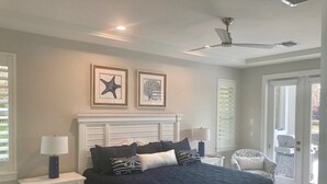 Une chambre à coucher comprenant un lit, des tables de chevet, un ventilateur de plafond et deux œuvres d’art encadrées accrochées au mur.