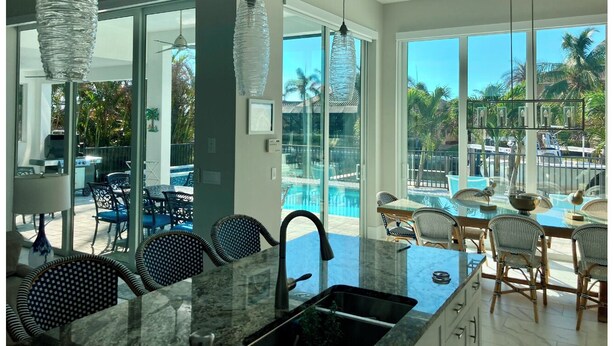 Une cuisine moderne dotée d’un grand îlot central, d’un coin repas avec des chaises en osier et d’une vue sur une piscine et des palmiers.