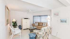 Un soggiorno con soffitto bianco, una ventola a soffitto, un divano beige con cuscini decorati, una poltrona bianca in vimini, un tavolino da caffè in vetro e una poltrona con motivi floreali.