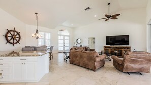 A spacious living area with a large TV, comfortable seating, a kitchen island, and a ceiling fan.