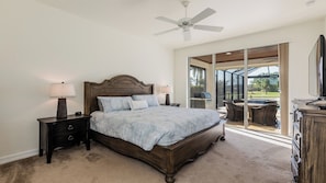 A bedroom with a large bed, two bedside tables with lamps, a ceiling fan, and a sliding glass door leading to an outdoor patio with a grill and seating area.