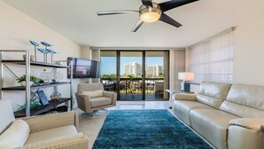 Ein modernes Wohnzimmer mit Deckenventilator, Flachbildfernseher, Balkon mit Blick auf Gebäude und einem blauen Teppich.