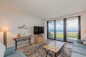 Relax in this stylish living room with a flat-screen TV, round table, and comfortable sofa, offering a view of a serene grassy area and water.