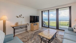 Ein Wohnzimmer mit Fernseher, Tisch und einer Couch. Ein Balkon bietet Blick auf den Strand.