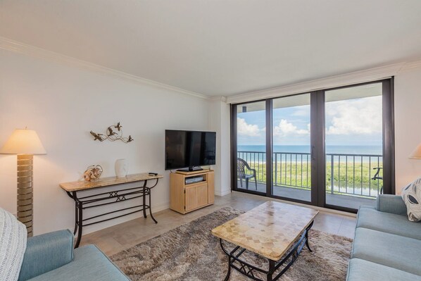 A living room with a TV, a table, and a sofa. There's a balcony with a view of the beach.