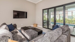 Una sala de estar moderna con sofás grises, un televisor de pantalla plana, una mesa de centro y un ventanal corredero que da a un balcón con piscina.