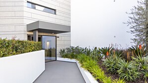 Un edificio moderno con una facciata pulita e geometrica, un vialetto stretto che conduce all'ingresso e diverse piante e arbusti sullo sfondo.