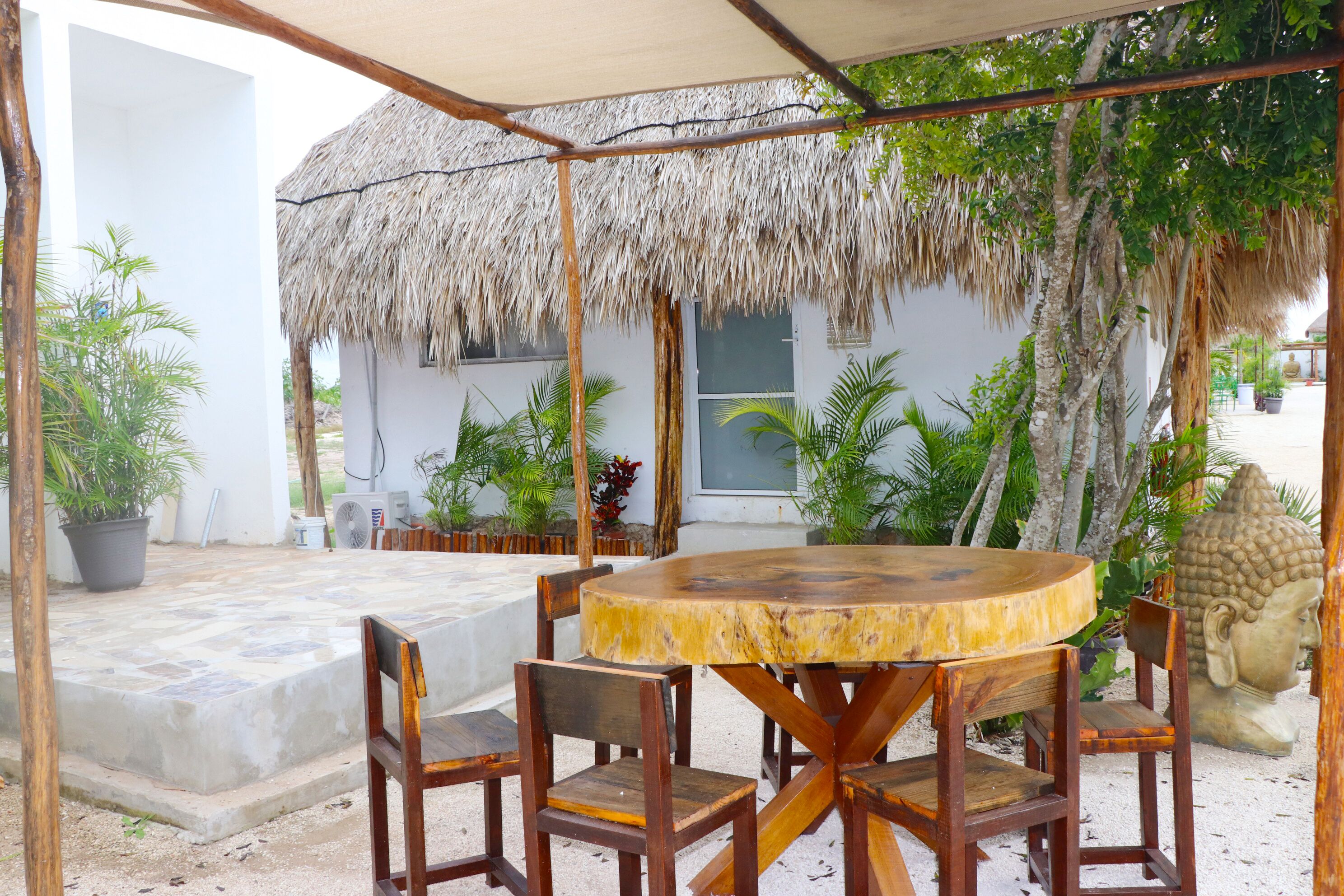 A round wooden table with chairs, a thatched roof, and a Buddha statue in a tropical setting.