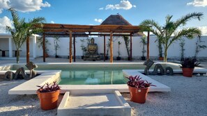 A serene outdoor pool area with a Buddha statue, a thatched-roof gazebo, and tropical plants.