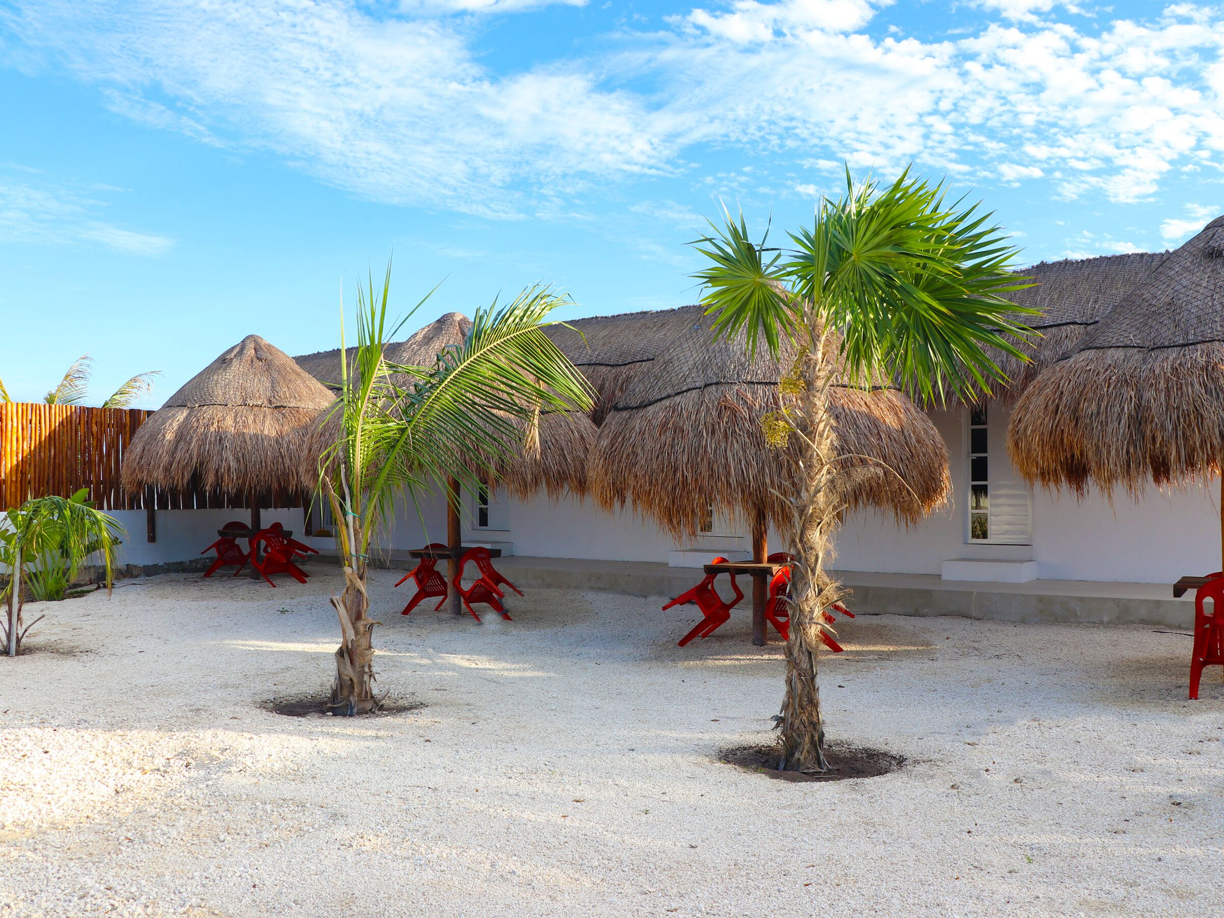 A courtyard with thatched-roof huts, palm trees, and red plastic chairs.