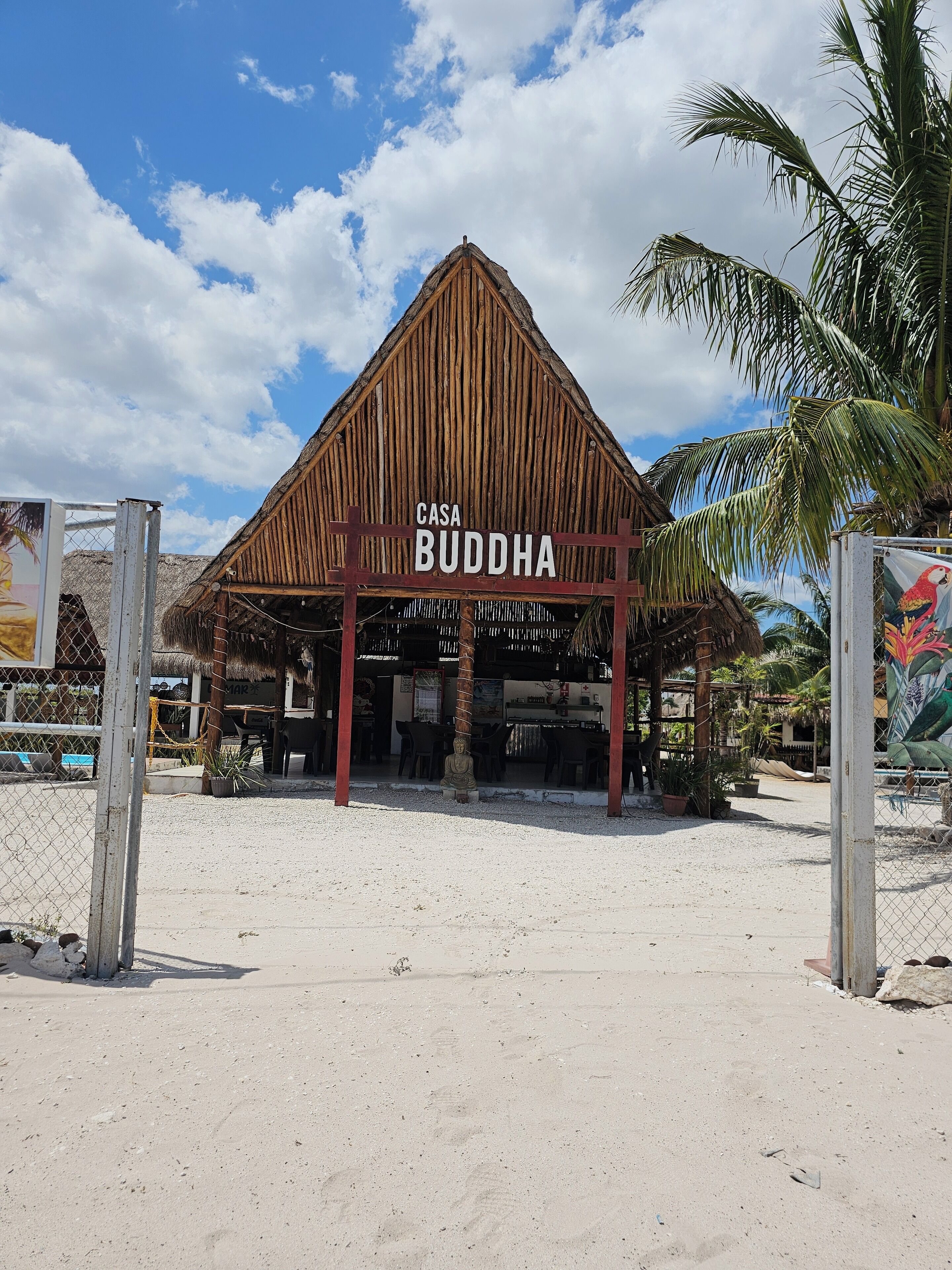 A thatched-roof structure labeled "Casa Buddha" with a sandy ground and palm trees.