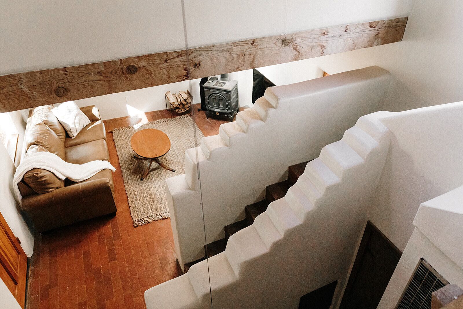 Un espacio habitable tipo loft con un sofá de cuero marrón, una mesa de centro redonda de madera y una radio antigua. Una escalera blanca conduce a un nivel superior.
