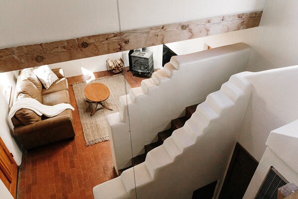 Um espaço de estar estilo loft com um sofá de couro marrom, uma mesa de centro redonda de madeira e um rádio antigo. Uma escada branca leva a um andar superior.