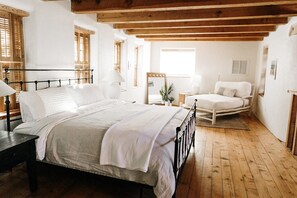 Quarto com piso de madeira, cama com roupa de cama branca, uma cadeira e mesa de cabeceira.