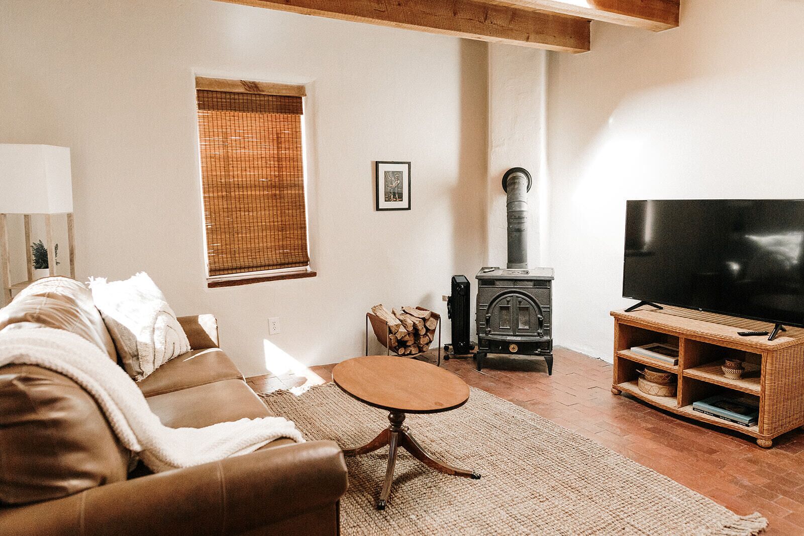 A cozy living room with a brown leather sofa, a round wooden coffee table, a television on a wooden stand, a wood-burning stove, and a window with blinds.