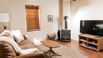 A cozy living room with a brown leather sofa, a round wooden coffee table, a television on a wooden stand, a wood-burning stove, and a window with blinds.