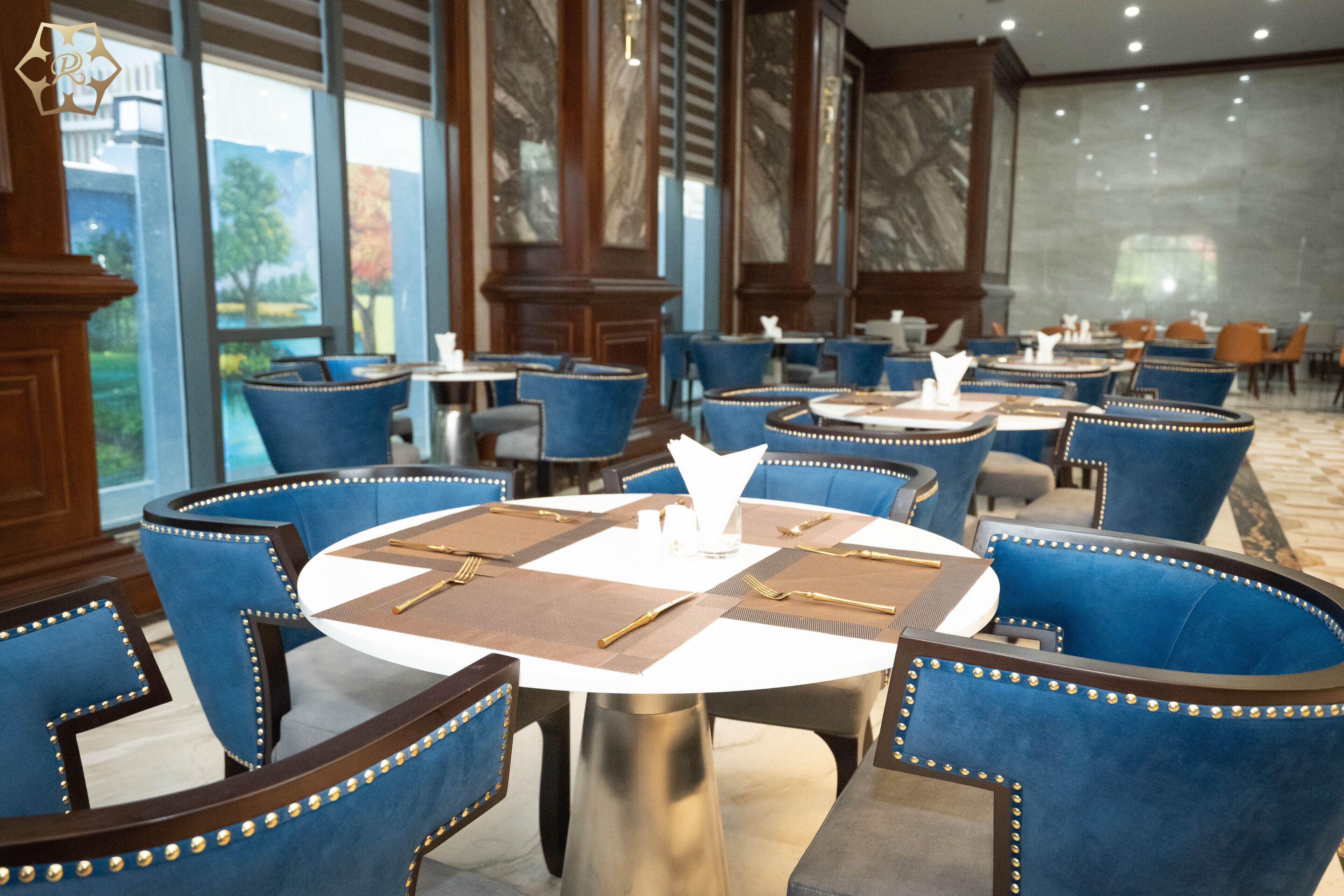 A dining area with round tables and blue upholstered chairs, featuring a marble wall and large windows.