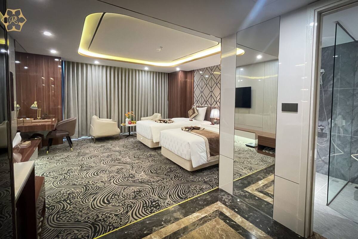 Une chambre d’hôtel comprenant un lit, deux tables de chevet, un bureau, une chaise, un espace douche et une moquette à motifs.