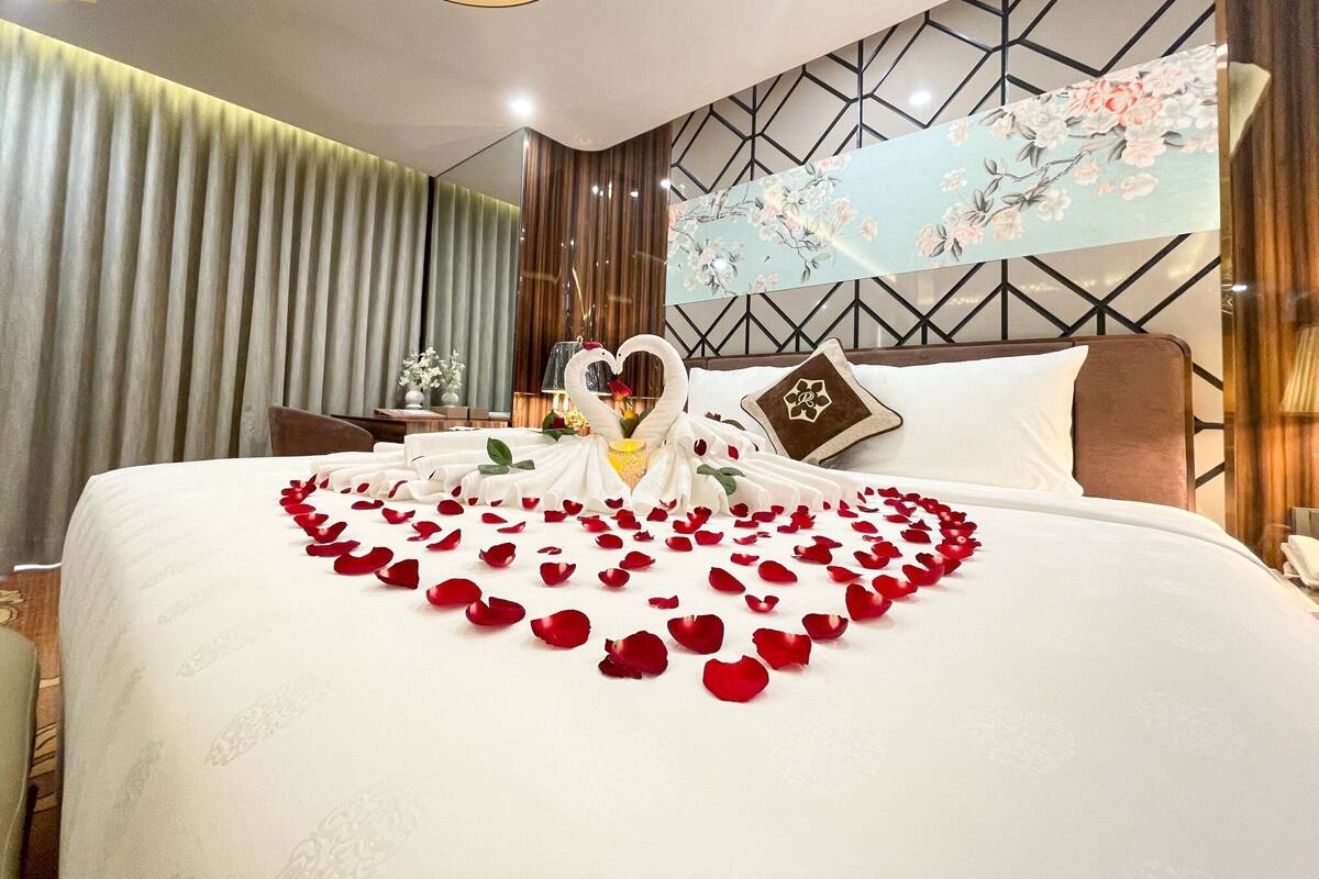 Une chambre d’hôtel avec un lit décoré de pétales de roses et d’une décoration en forme de cygne, une tapisserie murale à motifs floraux et un motif géométrique moderne sur le mur.