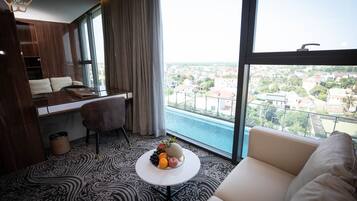 Habitació d'hotel moderna amb un sofà, una taula petita amb fruita, un escriptori i vista a un paisatge urbà a través de grans finestres.