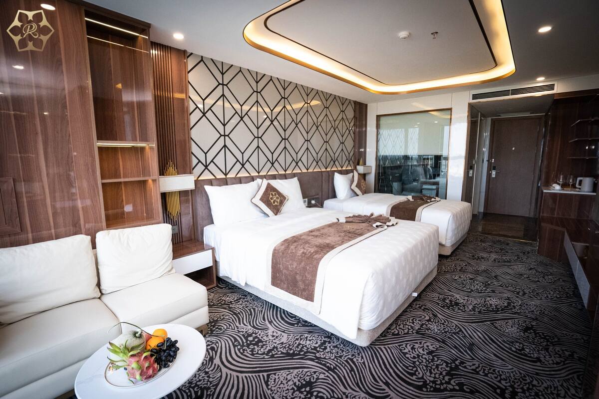 Une chambre d’hôtel moderne avec un grand lit, deux tables de chevet, un canapé et une petite table ronde sur laquelle sont disposés des fruits.