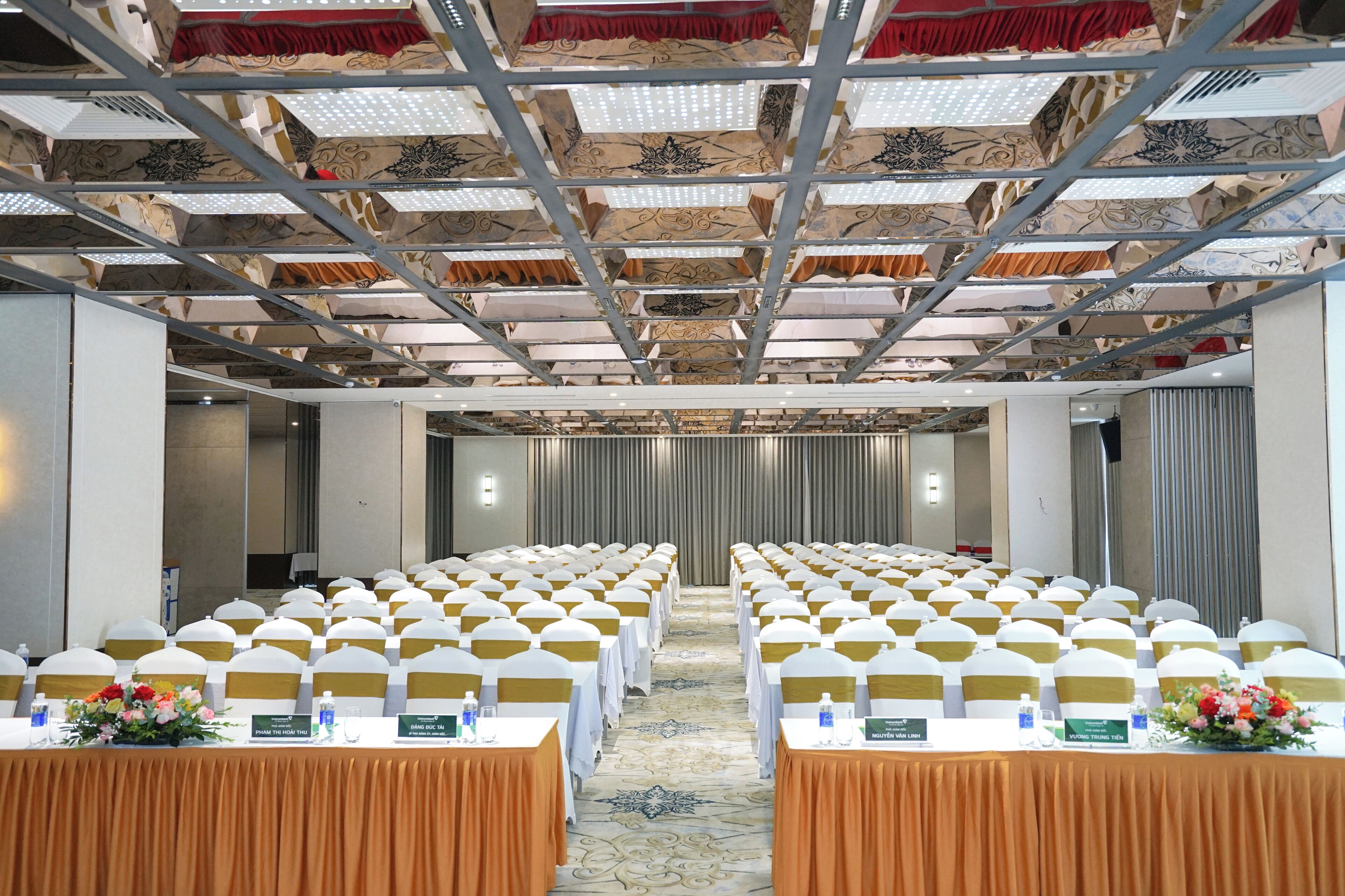 A conference room with rows of tables, chairs, and nameplates, featuring a patterned ceiling and a carpeted floor.
