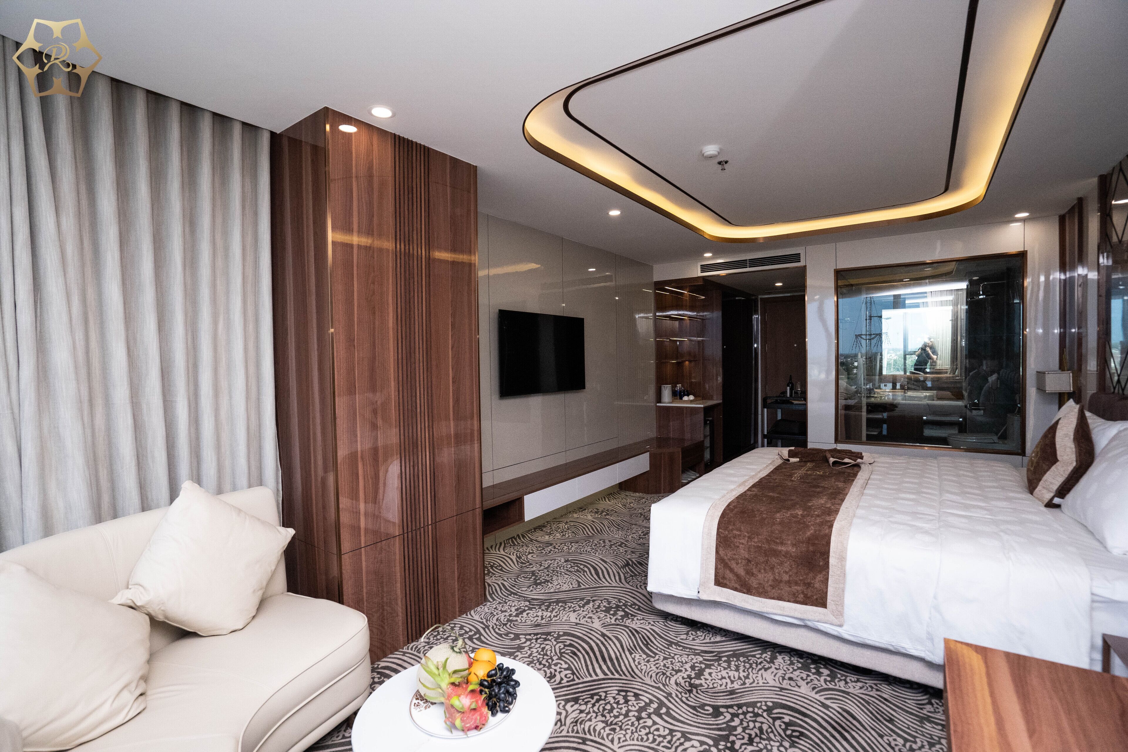 A modern hotel room with a sofa, a bed with a brown throw, a fruit bowl, and a TV on the wall.