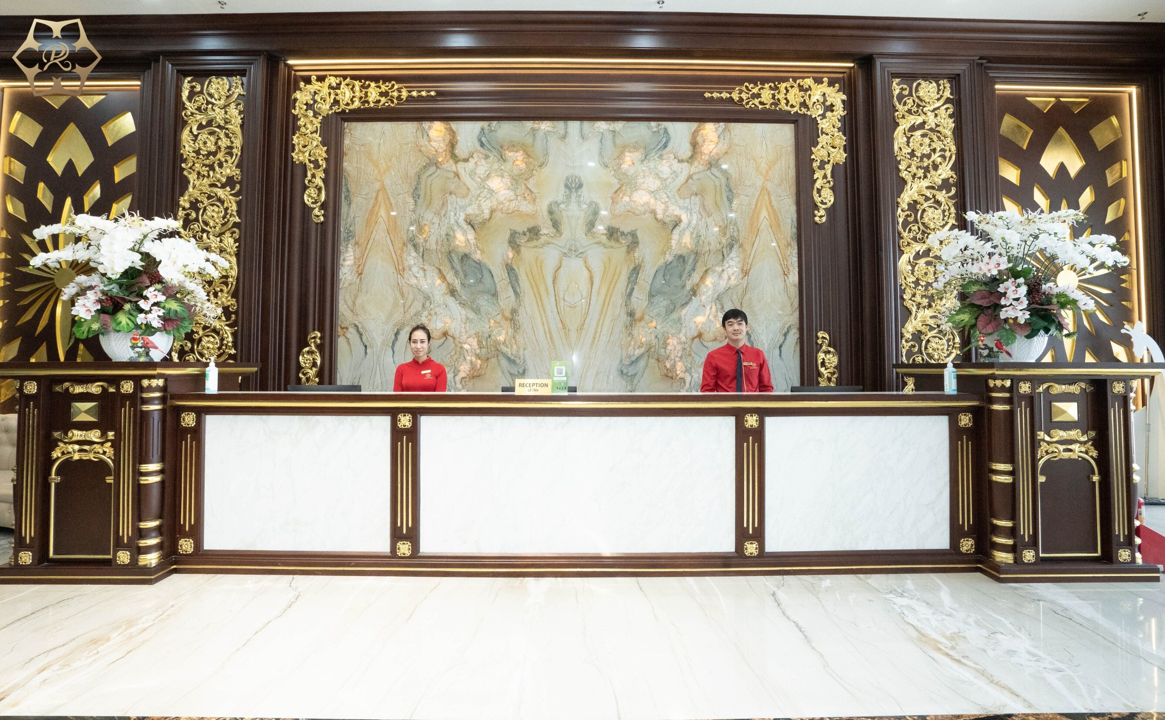 Un mostrador de recepción con dos empleados, paredes de mármol y detalles dorados decorativos.