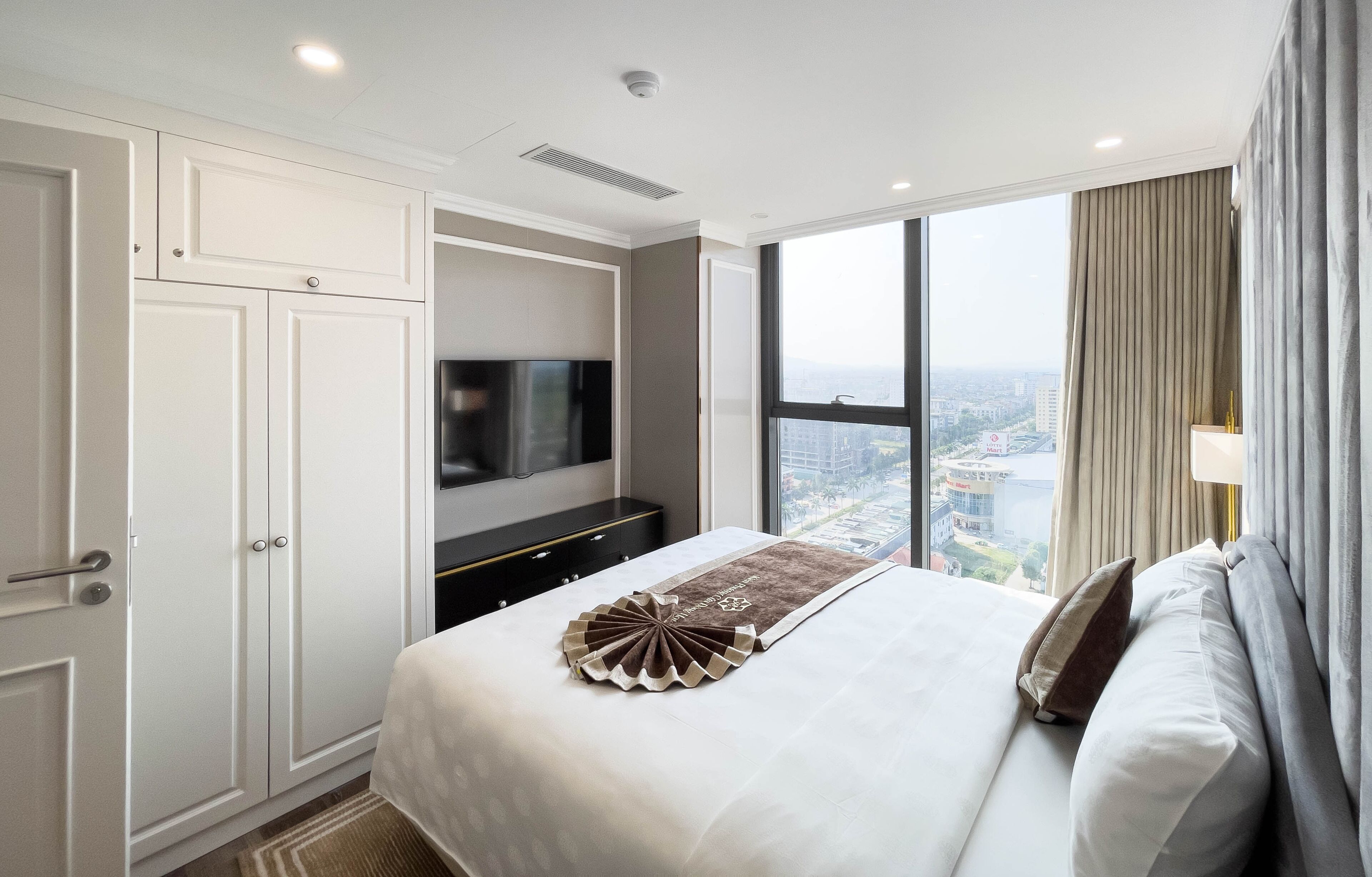 A hotel room with a large bed, a TV mounted on the wall, and a view of the city through large windows.