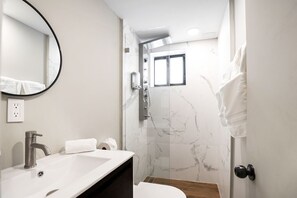 Une salle de bain moderne avec un lavabo blanc, un miroir, une douche avec une porte en verre et une petite fenêtre.