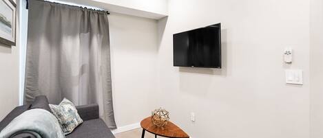 A modern living room with a grey sofa, a small wooden side table, a flat-screen TV mounted on the wall, and a window with grey curtains.