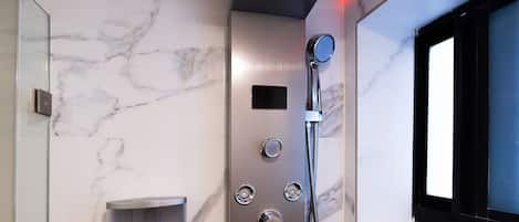 Un baño moderno con pared de mármol, ducha empotrada con múltiples mandos y una ventana con marco negro.