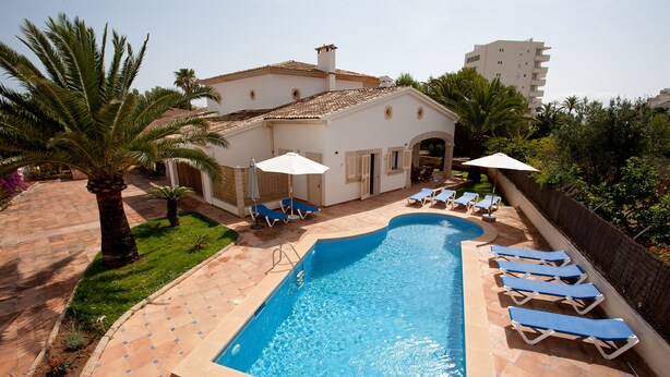 A swimming pool with lounge chairs and umbrellas, surrounded by palm trees and a building with a tiled roof. - LLAMPUDOL (VILLA SA COMA) - Wonderful villa with private pool and free WiFi. (Sant Llorenç des Cardassar)