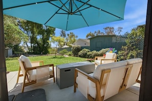 An outdoor patio with a table and chairs, a large umbrella, and a view of a backyard with a lawn and trees.