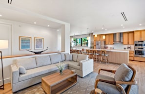 A modern living room with a sectional sofa, a wooden coffee table, and a patterned armchair. The kitchen features wooden cabinets, stainless steel appliances, and a bar area.