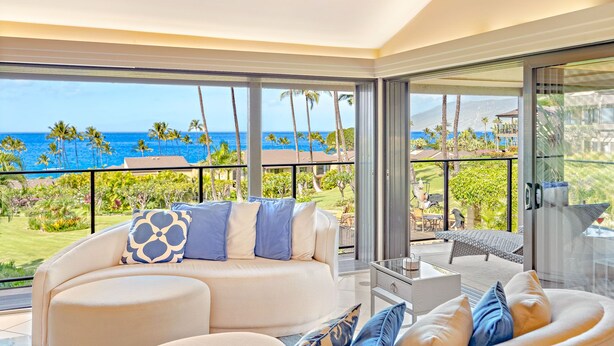 Ein modernes Wohnzimmer mit einer gebogenen Couch, einem Glastisch und Blick auf das Meer und Palmen.