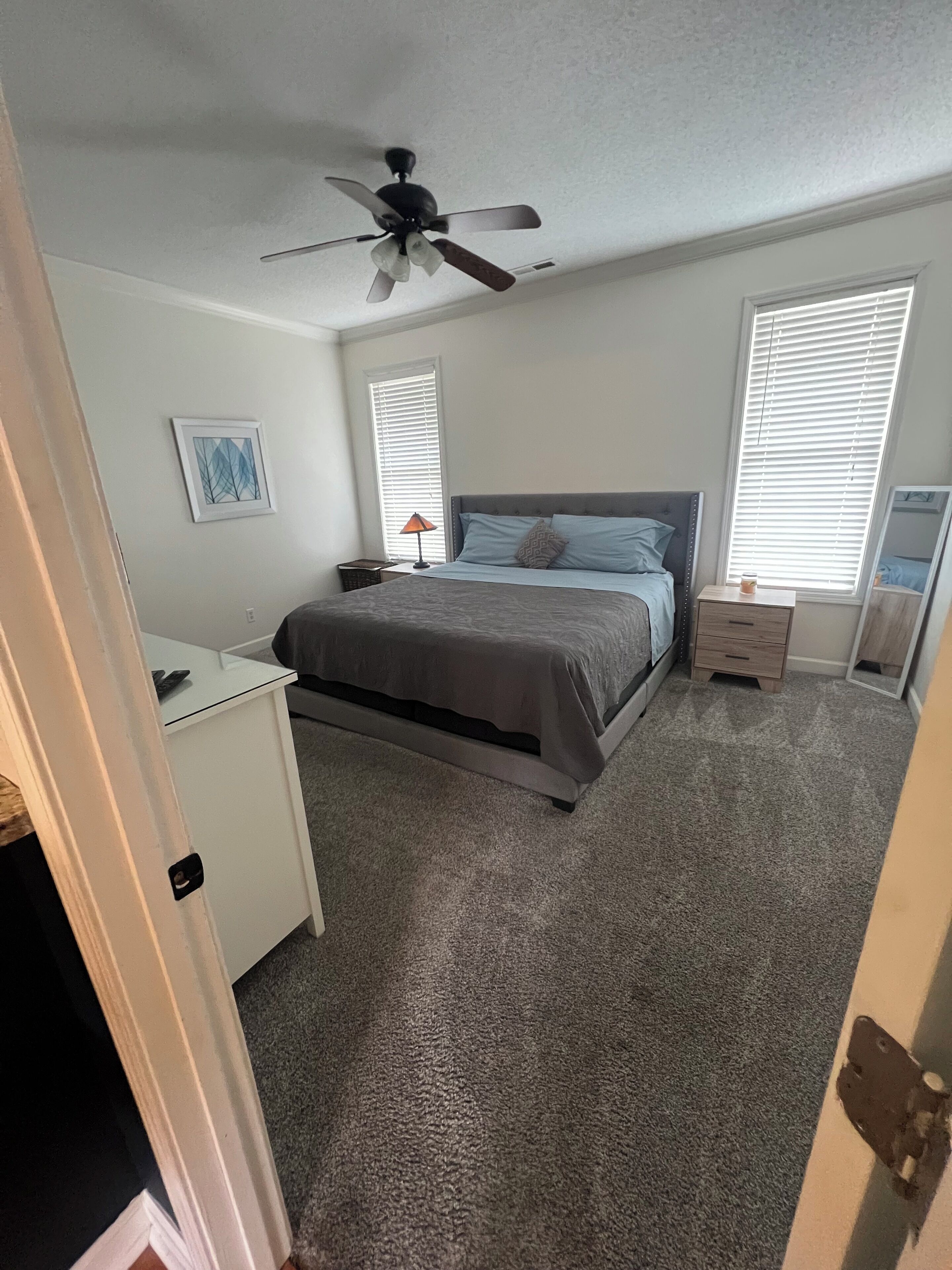 A bedroom with a bed, a desk, a ceiling fan, and two windows with blinds.