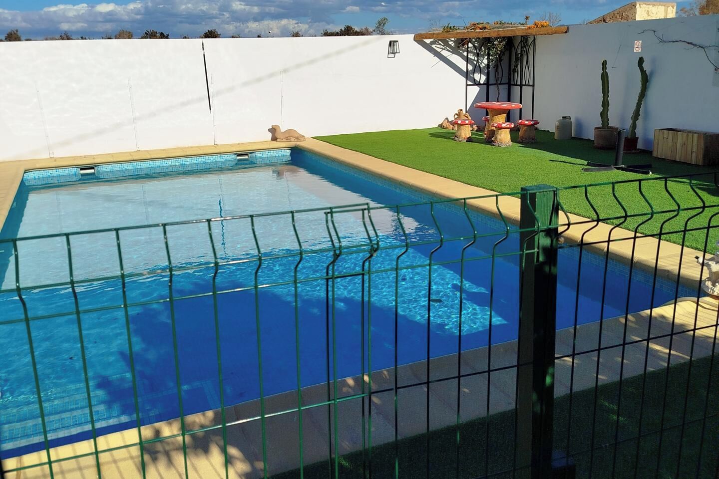 A rectangular swimming pool with a green fence, surrounded by a white wall and a small patio area with artificial grass and mushroom-shaped tables.
