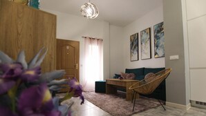 A modern living room with a wooden cabinet, a wicker chair, a coffee table, and a sofa. There are three framed artworks on the wall and a window with curtains. - RentalSevilla 3 bedroom apartment in Triana (Sevilla)