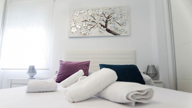 A neatly made bed with rolled towels, a white headboard, and a wall-mounted artwork of a tree. - RentalSevilla 3 bedroom apartment in Triana (Sevilla)