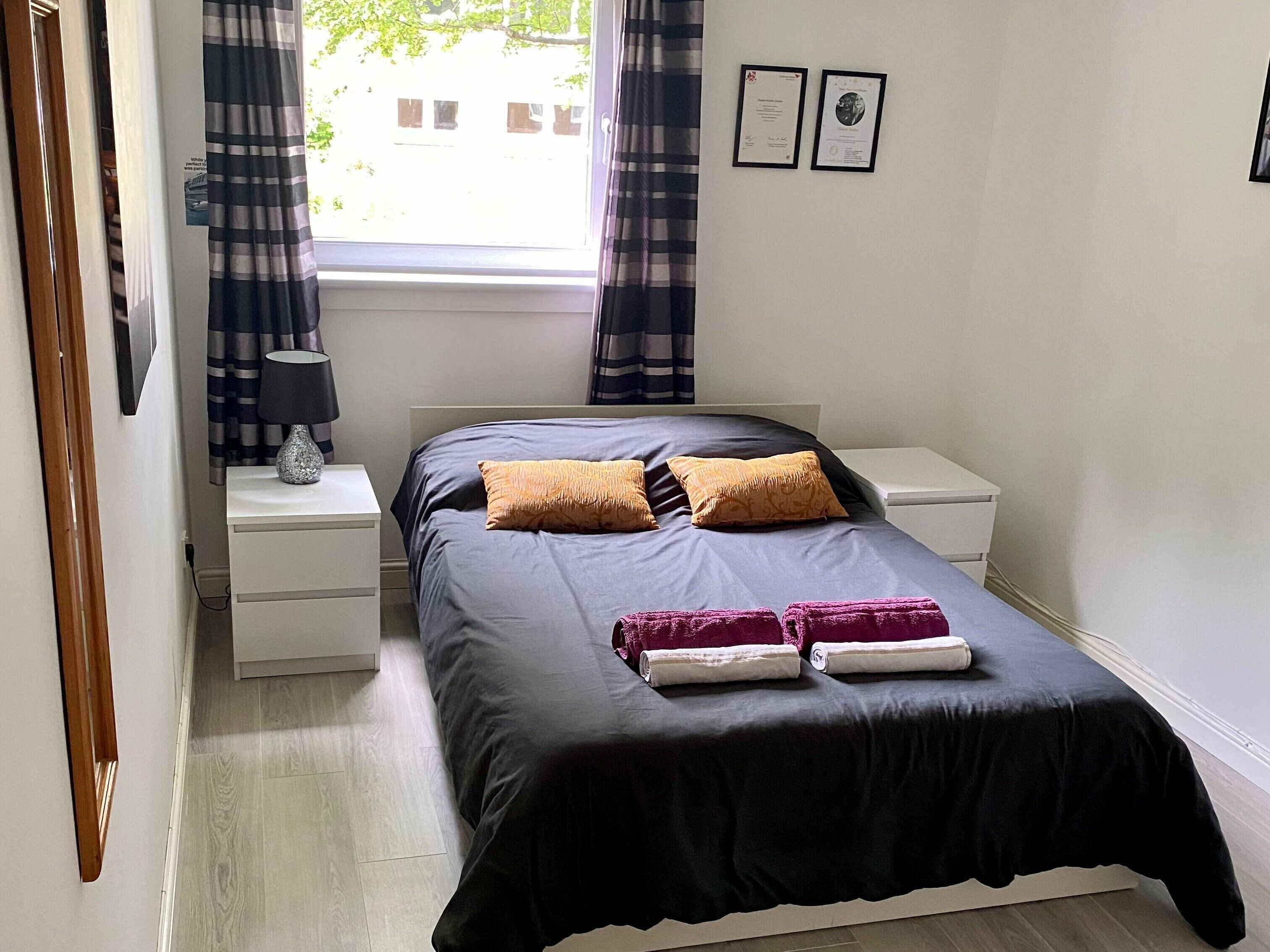 A neatly made bed with a black bedspread and two pillows, a bedside table with a lamp, and a window with curtains.