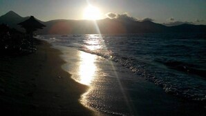 Ein Strand mit untergehender Sonne über dem Meer, deren Spiegelung sich im Wasser zeigt.